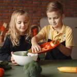 Little girl and boy mixing cutted pepper with other fresh ingredients for salad in big bowl while cooking at table with various fresh vegetables and fruits