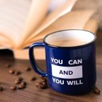 Inspirational coffee moment with a motivational quote on a mug beside an open book.